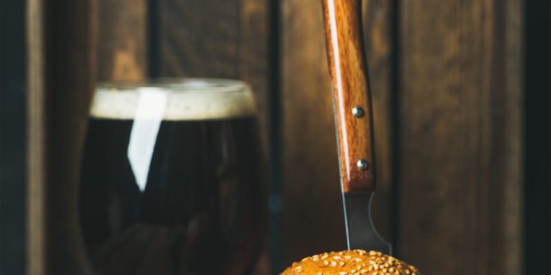 Homemade beef burger with crispy bacon, fresh vegetables and tomato sauce in cast iron pan served with glass of dark beer on rustic wooden background, selective focus, copy space, vertical composition