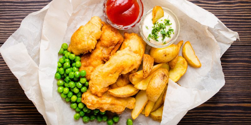 British national traditional fast food fish and chips with assorted dips, fresh peas, on paper, rustic brown wooden background, top view. Battered fried fish, potato chips, tartare and ketchup sauce